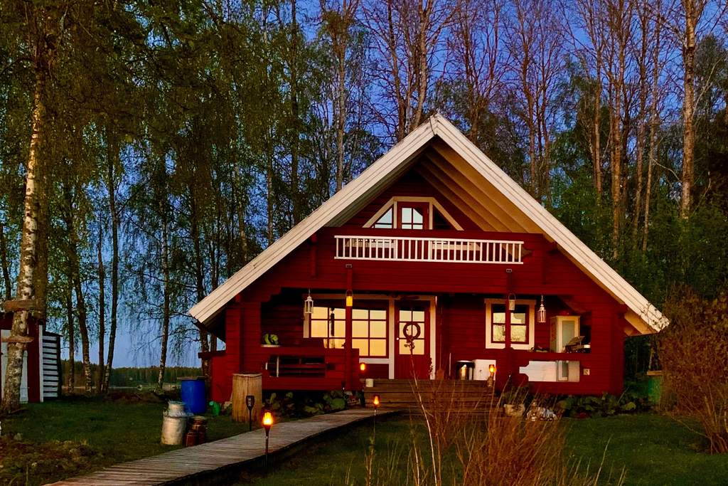 The red log house Aurinko at dusk, lit windows and a boardwalk through the grass